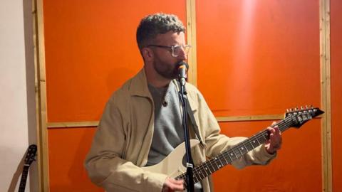 A standing man playing a guitar with a standing microphone close to his mouth. The man has short brown hair, a beard and is wearing glasses. He is wearing a beige coloured jacket on top of a grey top. He is stood in front of an orange wall. 