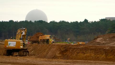 A yellow excavator and tipper truck excavate light brown sandy soil on a field, with the white dome of Sizewell B nuclear power station visible above tall trees on the horizon.