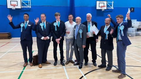 A group of men in suits stand in a sports hall. Some of the men have their arms raised in celebration.