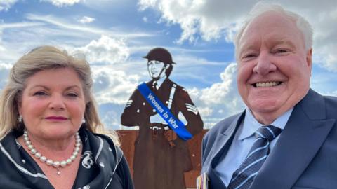 David Hourigan and his sister, Elizabeth, standing in front of a metal silhouette statue of a female soldier wearing a blue sash that reads "Women in War." Elizabeth stands on the left wearing a black blouse with a patterned collar, with a pearl necklace and pearl earrings. She has long blonde hair. David is standing on the right wearing a navy suit with a blue shirt and a dark and light blue striped tie. He is smiling at the camera and has short white hair. The sky is blue with light clouds. 