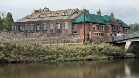 A general view of the Durham Baths building which sits in front of a bridge. The red-brick building has boarded-up windows and green netting on part of the roof.