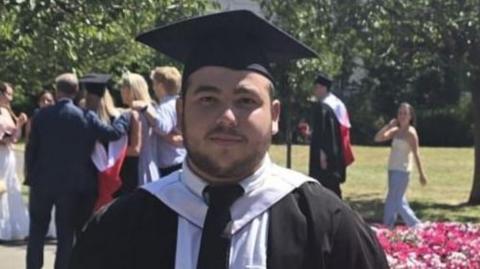Oliver, who has dark hair and short facial hair, is wearing graduation robes and a cap, standing beside a flower bed in a park, with other graduates in the background.