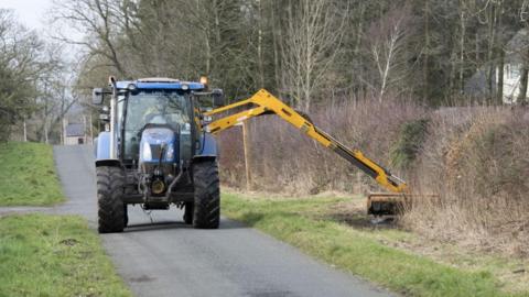 A tractor cuts grass verges along a rural road.