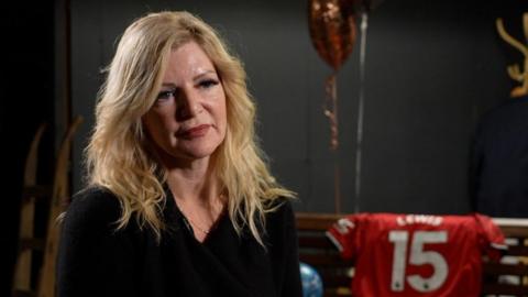 A woman with blonde hair and a black jumper sits in a room wih black walls, there is a red football shirt behind her which says 'Lewis 15' and some balloons. 