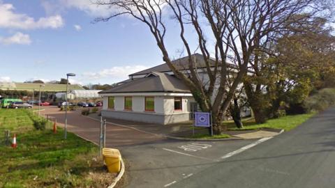 The exterior of Centre 21, which is a white building with a grey sloping roof. There are , with trees along the road next to it.