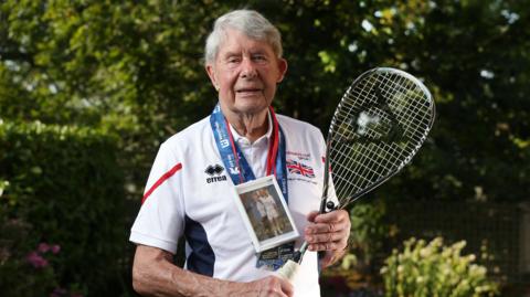 Mike Gibbons holding a squash racket and wearing a medal and a picture of him and his wife Ann round his neck 
