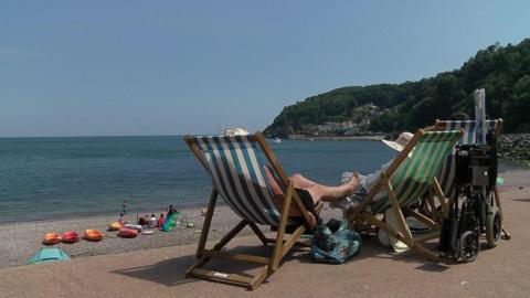 Two people relaxing in deck chairs overlooking a sandy beach. One person is resting their legs on the other person's chair. Calm sea water is lapping on the sandy shoreline. It appears to be a warm day. Four kayaks are visible on the beach in the background. 