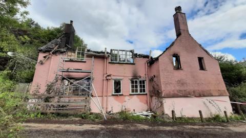 The remains of the destroyed home in the immediate aftermath of the fire. It is a large pink coloured home with two tall chimneys on either side. The exterior is covered in water and smoke damage, and the home is surrounded by greenery and trees. The roof of the building is completely gone and the inside is charred and smoking. 