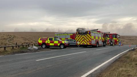 Fire vehicles parked on a road across a moor, with smoke rising in the background