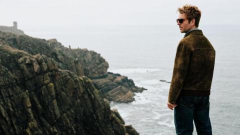A man looking out to the side with the sea in front of him and cliff paths to the left.