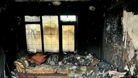 The inside of a living room which has been ravaged by fire. Huge chunks of paint and plaster are falling off the walls, the sofa is singed and the window is warped.