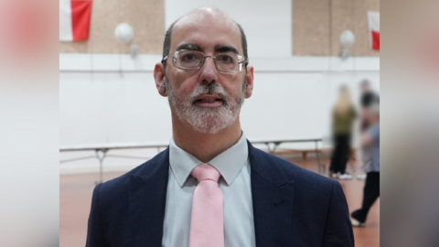 Anthony Felton is looking towards the camera in a school gymnasium with children blurred in the background. He is bald on top with close cropped dark hair on the sides. He is wearing glasses and has a trimmed white beard.