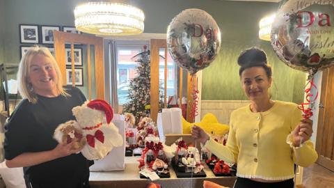 Two women stand by a table full of gifts for Christmas Eve gift boxes, including teddy bears, balloons and blankets. The woman on the left has bobbed blonde hair and a black t-shirt. The woman on the right has dark hair in a bun and wears a yellow cardigan, they are both smiling. 