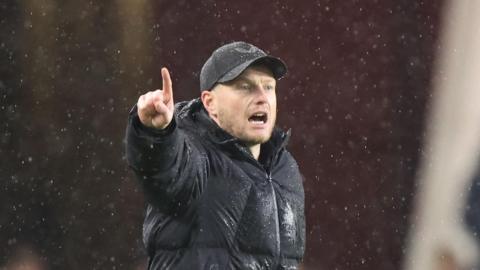 Kim Hellberg, wearing a cap and a large black puffer coat, points with right hand as the rain comes down during Middlesbrough's game with Derby