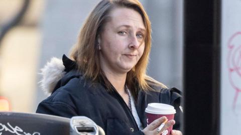 Woman carrying a coffee cup and cigarette, photographed from the side on a public street