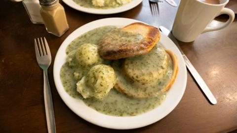 A picture of a white plate loaded with two pies, two balls of mashed potato and covered in traditional green liquor. The table top is brown and there is a knife and fork beside the plate as well as a cup of tea and salt and pepper shakers on the table.