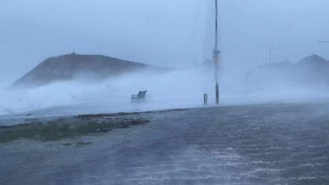 Waves at Troon harbour during Storm Eowyn. The picture is taken through a car window with the road in front of it completely flooded. Waves are crashing in the distance over a seating bench.