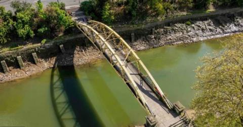 An aerial image of Langton Street Bridge before the repair work started. It is a yellow banana shaped metal bridge. The riverbanks are stripped back to mud and stones and appear somewhat unstable against rising tides.