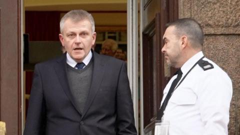 Deputy Philip Ozouf is walking out of a wooden door at Jersey's Royal Court building. He is wearing a dark suit and grey jumper and looking into the camera with his mouth open. A court usher in a black and white uniform is standing by the door.