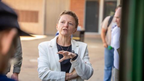 Manchester Mayor Bev Craig has short blonde hair and wears a white blazer. She is mid-conversation, standing outside addressing a people