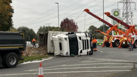 A white lorry on its side with orange cranes beside it and traffic cones