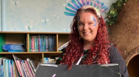 Teacher Lauren Noone with long red, curly hair and glittery make up. She is a holding a sign representing her word 'luminescent'