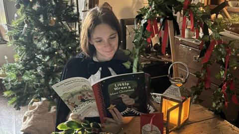 Author Felicity Wood sits at a table in front of her christmas tree whilst reading her new book 'Little Wren, Manx Christmas'.