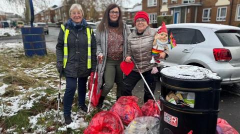 Three female volunteers are stood in a Blackburn street next to a bin surrounded with full bin bags. They are all dressed in winter clothing and are holding litter picker sticks. One woman is holding a garden gnome which is wearing rainbow shorts and holding a rainbow flag.