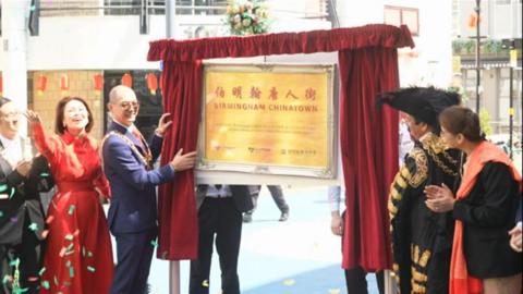 A large plaque reading "Birmingham Chinatown" stands in the street, with red curtains either side of it. Confetti is in the air, and people are clapping and smiling either side of the plaque