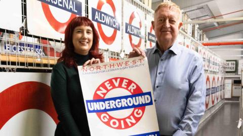 From left: Zoe Few and Ian Wilmott hold one of the original Never Going Underground posters used in the Manchester 1988 campaign against Section 28. Behind them is a wall of large Transport for London roundel signs.
