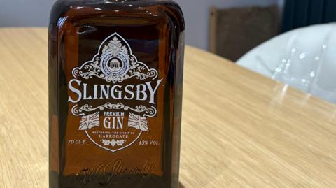Brown glass bottle is on a table. It reads "Slingsby Gin. Premium Gin. Restoring the Spirit of Harrogate."