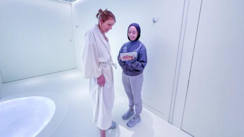 A person in a white dressing gown looks down at a tablet someone in trousers and a blue jacket is holding towards them. They are standing in a white room. 