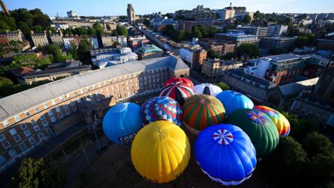 A group of 11 colourful hot air balloons are seen from above. They are fully inflated on College Green in Bristol as a publicity event for the 2026 Bristol International Balloon Fiesta. In the background Park Street is visible leading uphill towards the Wills Memorial Building, a large stone tower, in the distance