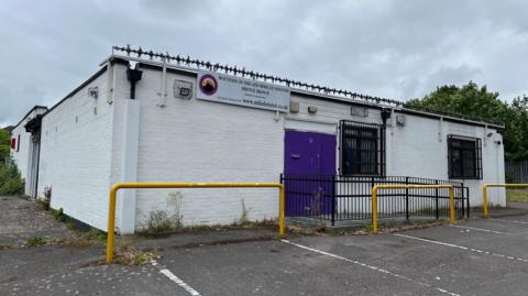 The single-storey brick building has been painted white. It has a purple door and bars on the windows. It stands in a car park and has barbed wire on the roof.