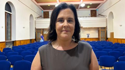 Gemma Brolly stands in Coleraine Town Hall after the meeting with rows of empty blue chairs behind her. She has shoulder length dark hair and is wearing a sleeveless brown top.
