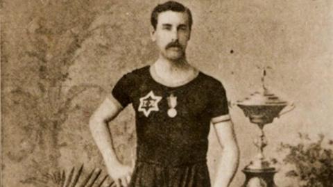 Sepia-coloured photograph of Charles Bennett standing next to a large trophy. He has dark hair and a large moustache and is wearing a dark t-shirt with a medal on the front