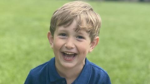 Benedict, a young boy with blond hair, is on a lawned space. He is wearing a blue polo shirt and is laughing into the camera.