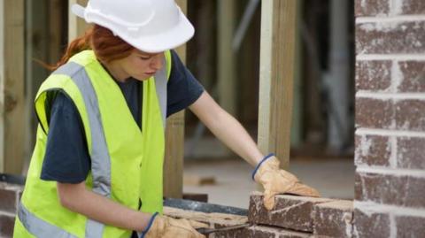 A woman wearing a high-vis jacket and hard hat laying bricks