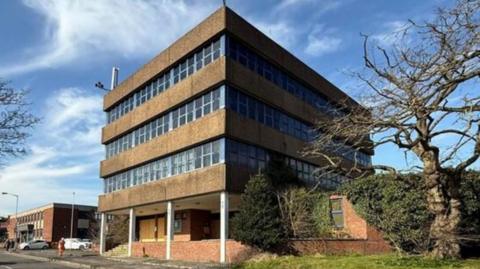A large four-storey building with alternate layers of brown concrete and glass windows with four white support pillars on the ground floor and a road running at the front.
