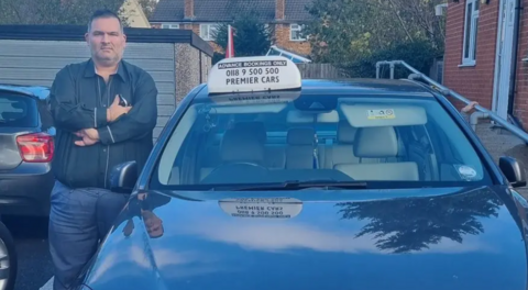 Kamran Saddiq standing crossed armed and frowning next to his dark car, wearing a navy shirt and blue trousers. 