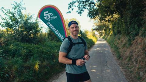 A man wearing running gear standing on a small country lane with a sign behind which says Les Amis 50 on it and the hashtag miles4smiles.