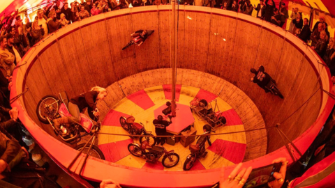 Bikers ride round a wall of death - a mostly wooden structure. Crowds have gathered around it. Three people are riding bikes on the sides of the wall of death while a man is stood at the bottom.