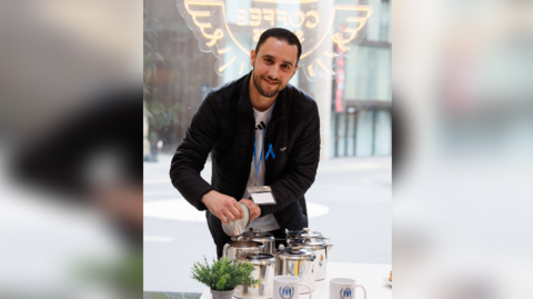 Smiling Mouhyedin Alkhalil pouring tea into silver pots on a table with a green plant in the foreground