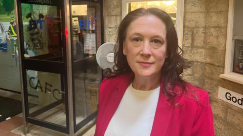 A woman in a red blazer and white top stands indoors near a vintage glass telephone booth with “Telephone” signs and the old BT logo. The setting includes a stone-textured wall, a small lit window, and part of a sign reading “God.”