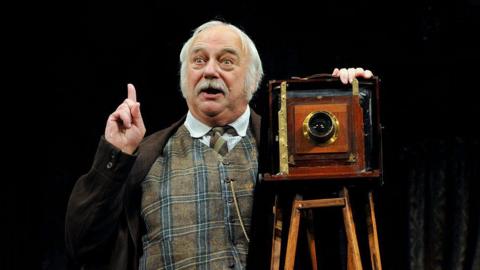 Roy Hudd as Henry Ormanroyd in the production of JB Priestley's When We Are Married at London's Garrick Theatre. He is wearing a tweed waistcoat, tie and a brown pinstripe jacket. He has his left hand resting on an old Victorian camera, and his right index finger is pointing upwards.