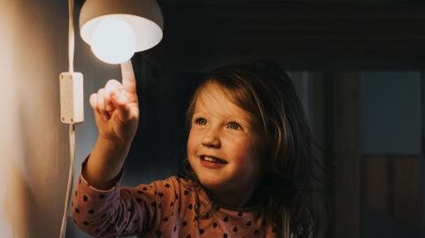 A young girl wearing pyjamas touching a light blub in a lamp with her index finger 