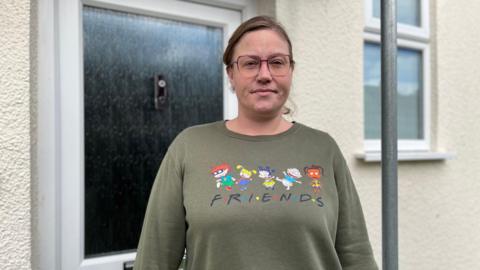 A woman with brown hair, wearing glasses with a diamond stud under her nose. She's wearing a green jumper with the word Friends on it and the characters from the Rugrats. 