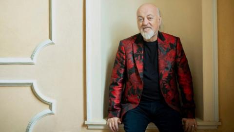 Comedian Bill Bailey wears a red floral blazer, black t-shirt and trousers. He sits leaning against a cream coloured wall and smiling. 