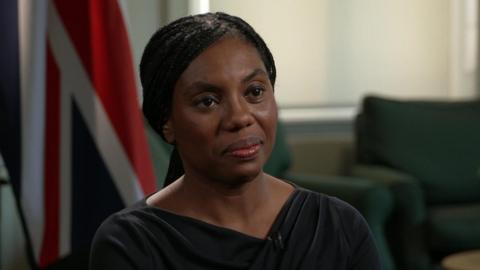 Kemi Badenoch, wearing a black dress, sits in a room. There are green armchairs and a union flag visible in the background. 