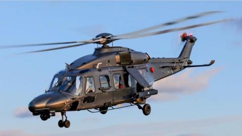 A photograph of an AW149 flying. There is a red tail light on and the wheels are down. 